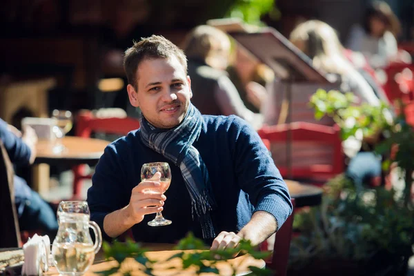 Joven sentado en la mesa de bar al aire libre tomando una copa de vino blanco y mirando a la cámara — Foto de Stock