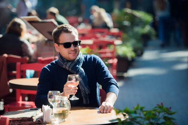 Joven sentado en la mesa de bar al aire libre tomando una copa de vino blanco y mirando a la cámara — Foto de Stock