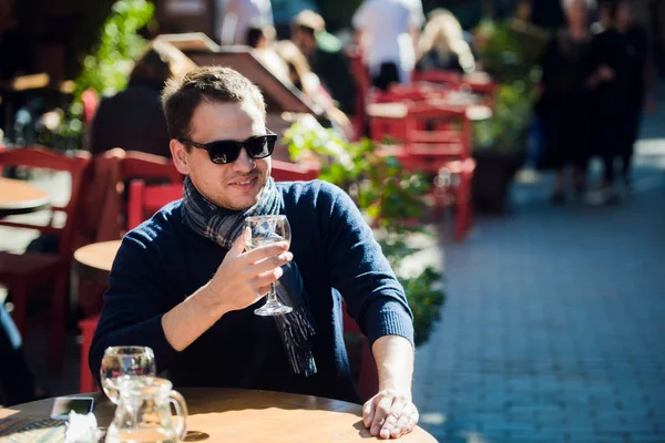 Joven sentado en la mesa de bar al aire libre tomando una copa de vino blanco y mirando a la cámara — Foto de Stock