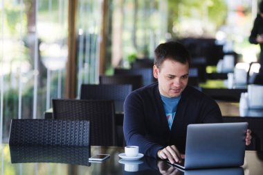 oturma ve kafede dizüstü bilgisayar ile eğitim genç erkek öğrenci konsantre. Büyük cam pencereler kapalı bir arka plan üzerinde