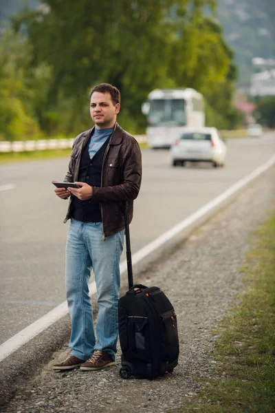 Man standing on the road with suitcase and tablet pc waiting for car