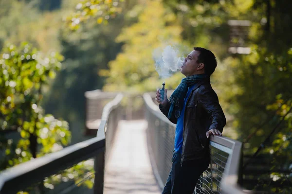 Νεαρός άνδρας με προσαρμοσμένο πλαίσιο λειτουργίας vape συσκευή ατμίσματος ένα ηλεκτρονικό τσιγάρο που είναι εξωτερική στο όμορφο μέρος σε εξωτερικούς χώρους — Φωτογραφία Αρχείου