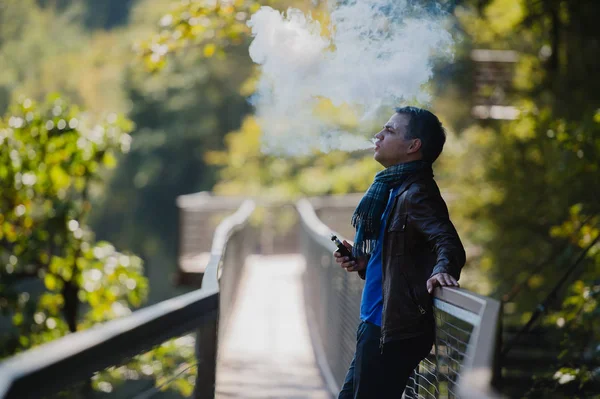 Ung man med anpassat läge vape enhet vaping en elektronisk cigarett utomhus på vacker plats utomhus — Stockfoto