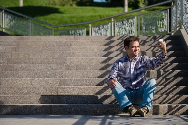 Jonge, gelukkige man zittend op de trap, het bezit van mobiele telefoon en het maken van een selfie. — Stockfoto