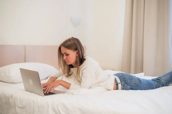 Mujer rubia atractiva pensativa usando una computadora portátil mientras se sienta en el borde de una cama en casa o en un dormitorio de hotel en vacaciones o viaje de negocios —  Fotos de Stock