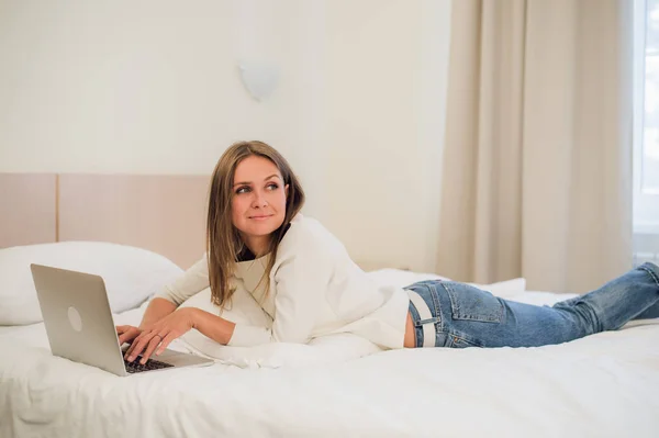 Mujer rubia atractiva pensativa usando una computadora portátil mientras se sienta en el borde de una cama en casa o en un dormitorio de hotel en vacaciones o viaje de negocios — Foto de Stock