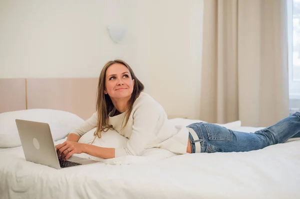 Una joven linda sonriendo emocionada en la pantalla de su computadora portátil. Ella se está relajando en la cama . — Foto de Stock