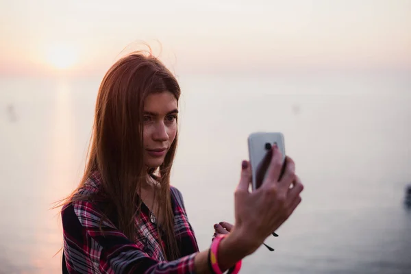 Портрет молодой счастливой женщины в хипстерской рубашке, стоящей перед живописным видом на море, делающей селфи с закатом или восходом солнца на смартфоне — стоковое фото