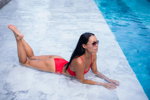 Femme sexy posant et détendue près de la piscine à cool noir lunettes de soleil à la mode, soutien-gorge casseroles de bikini, bronzage femme peau rougeoyante, coiffure étonnante, coiffure longue — Photo