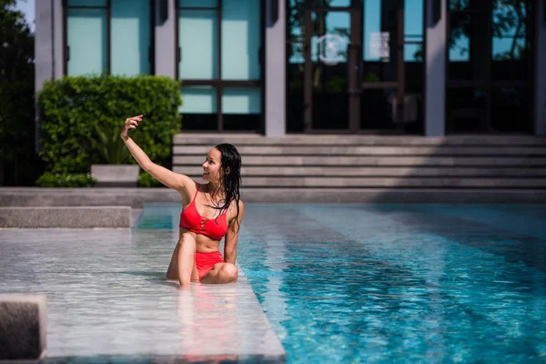 Personas, tecnología, viajes, turismo y concepto de verano - mujer joven feliz en bikini traje de baño y gafas de sol tomando selfie con smatphone sobre la piscina en la playa con palmeras fondo — Foto de Stock