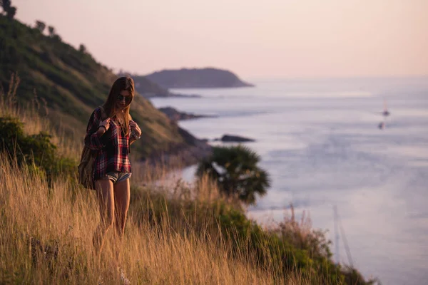 Летний солнечный образ жизни модный портрет молодой стильной хипстерши, гуляющей в горах, одетой в милую модную одежду, улыбающейся, наслаждающейся выходными, путешествующей с рюкзаком. Вид на океан — стоковое фото