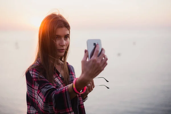 Портрет молодой счастливой женщины в хипстерской рубашке, стоящей перед живописным видом на море, делающей селфи с закатом или восходом солнца на смартфоне — стоковое фото