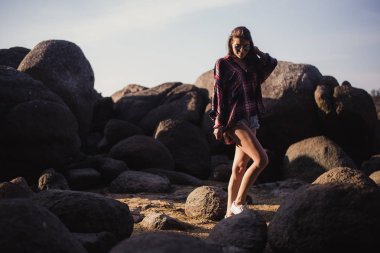 Güzel ince kadın gömlek, şort ve moda güneş gözlüğü büyük kayalar üzerinde yürümek. Arkadan görüntüleyin. Güzellik büyük taşlarla bir tropikal plaj deniz okyanus kıyısında şirin kız. Açık yaz yaşam tarzı.