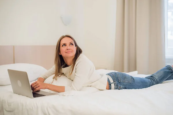 Una joven linda sonriendo emocionada en la pantalla de su computadora portátil. Ella se está relajando en la cama . —  Fotos de Stock