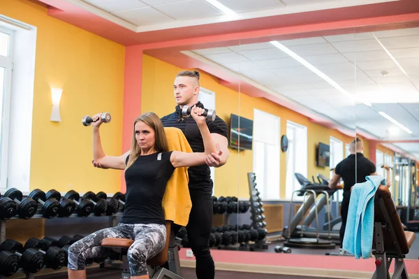 Allenamento in palestra: Bella donna yong che fa esercizi con i manubri seduta sulla panchina mentre l'allenatore muscolare la guarda e la assiste — Foto Stock