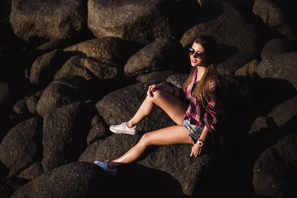 Sexy kobiety w dżinsach ogólnej i różowy t-shirt siedzieć na wielki kamień skały plaża morze ocean shore. Hipster nowoczesna dziewczyna zabawy. Dobry słoneczny letni dzień — Zdjęcie stockowe