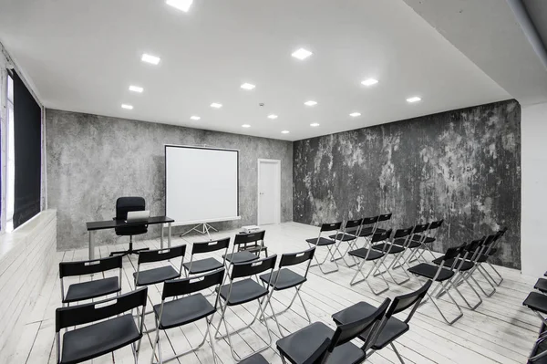 Room for lecture with a lot of dark chairs. Walls are white, loft interior. On the right there is a door. On the background there is a table with a laptop. — Stock Photo, Image
