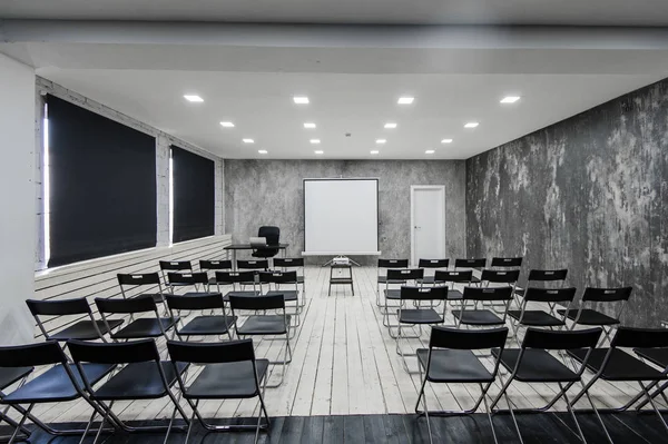 Room for lecture with a lot of dark chairs. Walls are white, loft interior. On the right there is a door. On the background there is a table with a laptop. — Stock Photo, Image