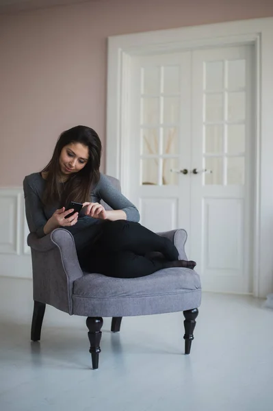 Chica sonriente relajándose en casa, se está conectando y las redes sociales con su teléfono inteligente — Foto de Stock