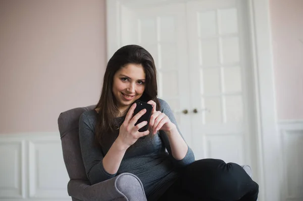 Chica sonriente relajándose en casa, se está conectando y las redes sociales con su teléfono inteligente — Foto de Stock