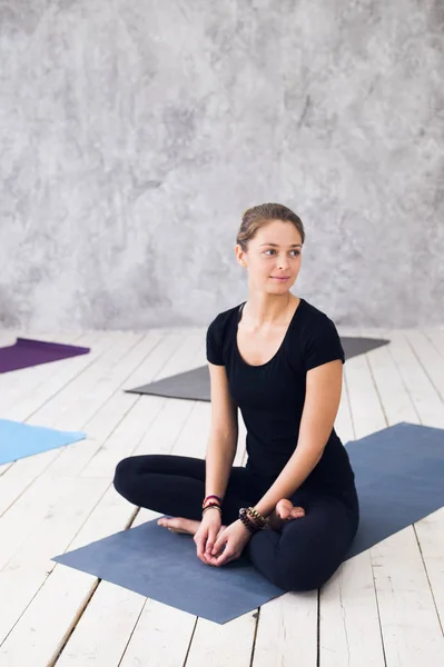 Ung attraktiv kvinna utövar yoga, sittande i Ardha Padmasana motion, halv Lotus pose, träna, klädd i svart t-shirt, byxor, meditation session på golvet vid yogaklass — Stockfoto