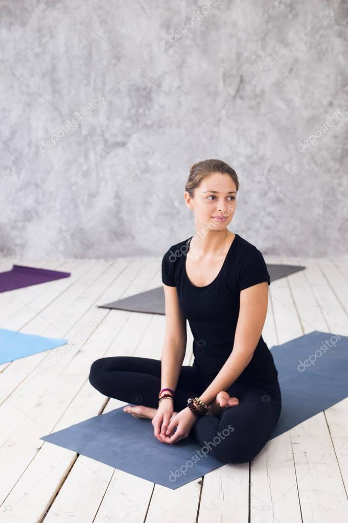 Young attractive woman practicing yoga, sitting in Ardha Padmasana exercise, Half Lotus pose, working out, wearing black t-shirt, pants, meditation session at floor at yoga class