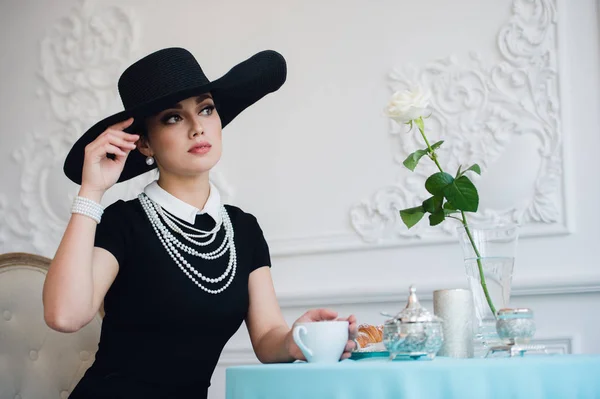 Kobieta w kapeluszu, jak słynna aktorka, rogalik, jedzenie i picie herbaty. — Zdjęcie stockowe