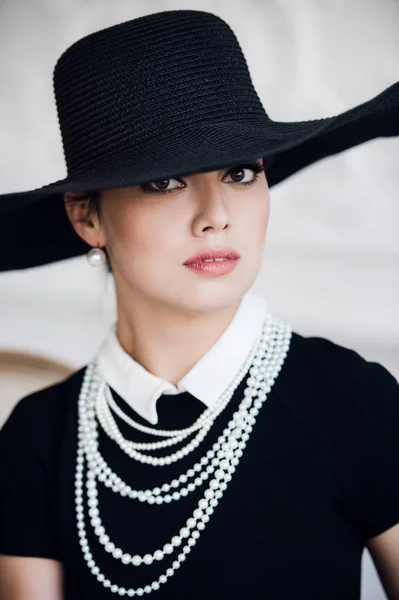 Atrakcyjna kobieta ubrana w czarną sukienkę, kapelusz i perły, siedzi na krześle — Zdjęcie stockowe