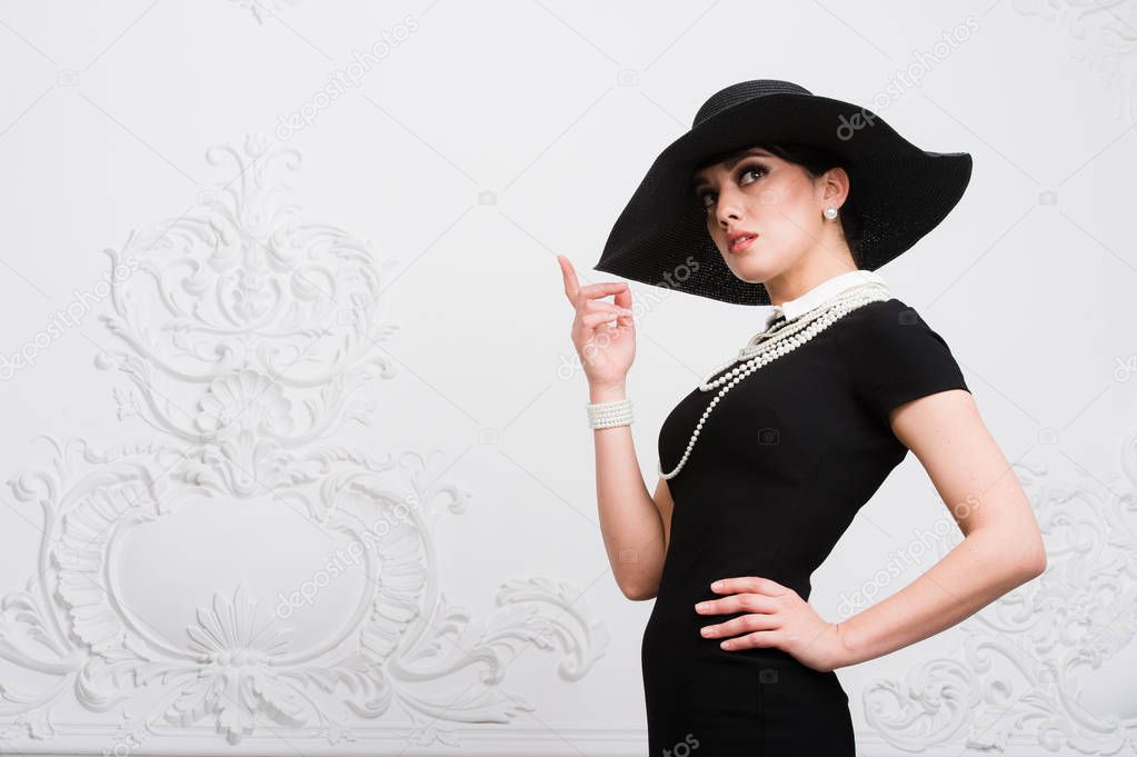 Portrait of a beautiful young woman in retro style in an elegant black hat and dress over luxury rococco wall background