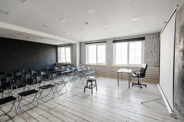 Room for lecture with a lot of dark chairs. Walls are white, loft interior. On the right there is a door. On the background there is a table with a laptop.