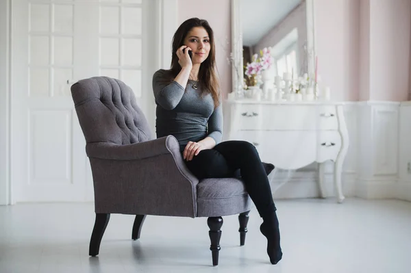 Fotografía de una mujer casual hablando con alguien en su teléfono móvil mientras está sentada en un sillón en casa . — Foto de Stock