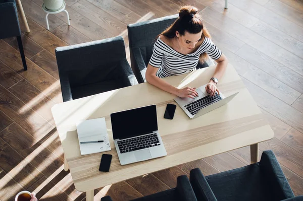 Visão geral da jovem mulher usando um smartwatch trabalhando em seu laptop em um café. Top vista tiro de fêmea sentado em uma mesa com um telefone nas proximidades e navegar na internet . — Fotografia de Stock