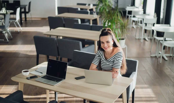 Joven hermosa mujer de negocios que trabaja en su computadora portátil en el interior, mujer adulta sonriente está utilizando un ordenador portátil en la acera cafetería, chica sonriente con computadora portátil en la cafetería . —  Fotos de Stock