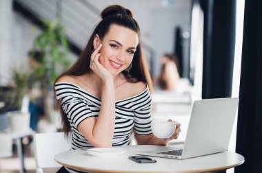 Gülümseyen ve kameraya bakarak ve kafede dizüstü kullanarak genç beyaz bir kadın.