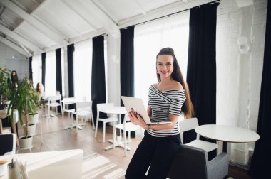 Gülümseyen ve kameraya bakarak ve kafede dizüstü kullanarak genç beyaz bir kadın.