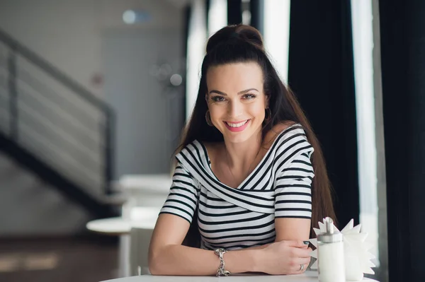 Mujer joven en blusa a rayas está sentado en la cafetería en la mesa de madera cerca de la ventana y mirando a la cámara. Sonrisa perfecta, interior moderno, interior . — Foto de Stock