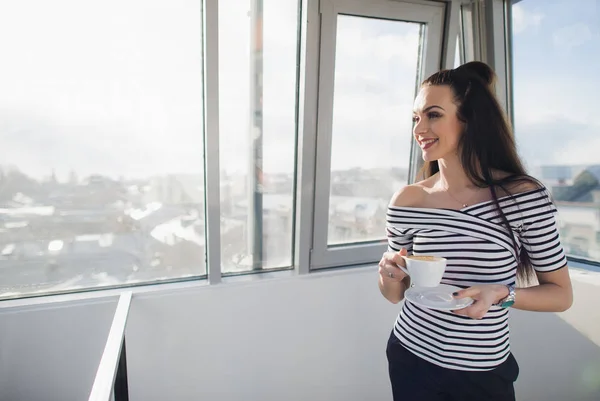 Retrato de una hermosa mujer sonriendo y mirando a través de la ventana sosteniendo una taza de té . — Foto de Stock