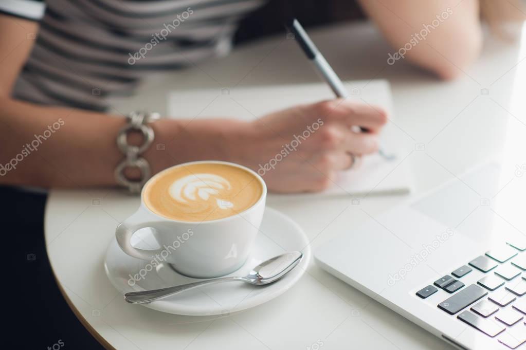 Close up picture of womans hands and a cup of cappuccino. Lady is writing in her notebook with a laptop nearby.