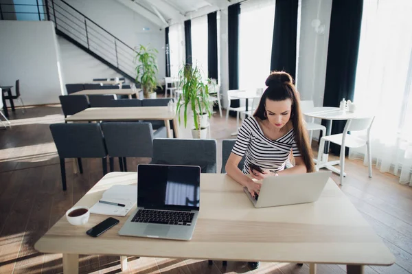 Jonge Kaukasische vrouw lachend en met behulp van laptop in café. Theres een andere laptop, telefoon en een kopje koffie in de buurt. — Stockfoto