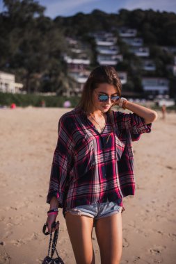 Yaz yaşam tarzı tropik ada sahile yürüme sportif tabaklanmış vücut, genç güzel kız portresi. Damalı t-shirt ve şık güneş gözlüğü takıyor. Küçük plaj çantası ile