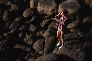 Güzel ince kadın gömlek, şort ve moda güneş gözlüğü büyük kayalar üzerinde yürümek. Arkadan görüntüleyin. Güzellik büyük taşlarla bir tropikal plaj deniz okyanus kıyısında şirin kız. Açık yaz yaşam tarzı.