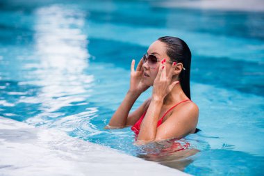 Islak saç dokunmadan kırmızı mayo ve yüzme havuzunda oturan güneş gözlüğü takan kadın