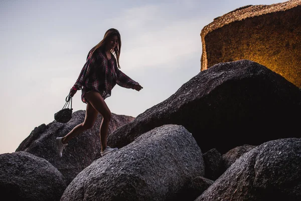 Schöne schlanke Frau in Hemd, Shorts und modischer Sonnenbrille spaziert auf großen Felsen. Blick von hinten. Schönheit nettes Mädchen an einem tropischen Strand Meer Ozeanküste mit großen Steinen. Sommerlicher Lebensstil im Freien. — Stockfoto