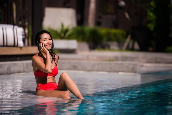 Llamando a todos sus amigos para unirse. Retrato de una joven feliz sentada junto a la piscina hablando en su teléfono riendo alegremente — Foto de Stock