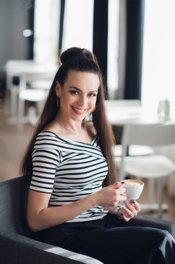 Bir kafede çay içme ile genç bir kadın. Bir fincan cappuccino bir kafede güzel yetişkin kadın. Sıcak içecek rahatlatıcı ile gülümseyen esmer.