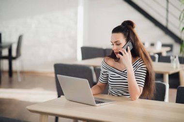 Güzel işkadını kafede sıcak kahve tadını çıkarırken bir telefon tutarak portresi. Bir dizüstü bilgisayar ile masada yetişkin çekici kadın oturma ve çalışma sorunları tartışıyor.