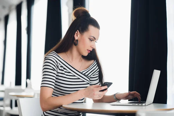 Portret van mooie zakenvrouw een telefoon te houden terwijl u geniet van warme koffie in het café. Volwassen aantrekkelijke vrouwelijke zitten aan de tafel met een laptop en bespreken werk kwesties. — Stockfoto