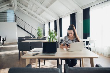 Hareket eden telefon tutarak ve açık dizüstü bilgisayar ile tahta masada otururken Sms tipik genç kadın.