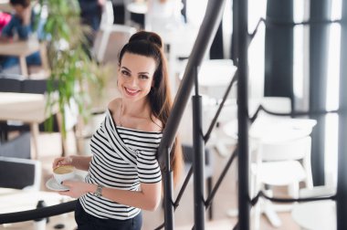 Bir fincan cappuccino bir kafede duran neşeli casual kadın.