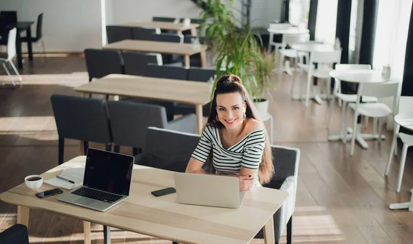 Volwassen aantrekkelijke vrouw zitten aan een tafel in een café met een laptop. Prachtige vrouwelijke kijken naar de camera en een bericht aan het typen. — Stockfoto